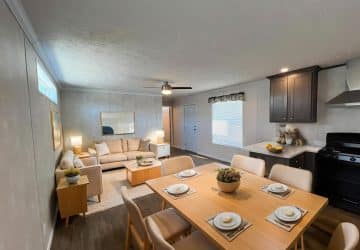 A modern dining and living area with a wooden dining table set for six, beige chairs, a cozy beige sofa, light wood furniture, and a kitchen with dark cabinets and a stove in the background.