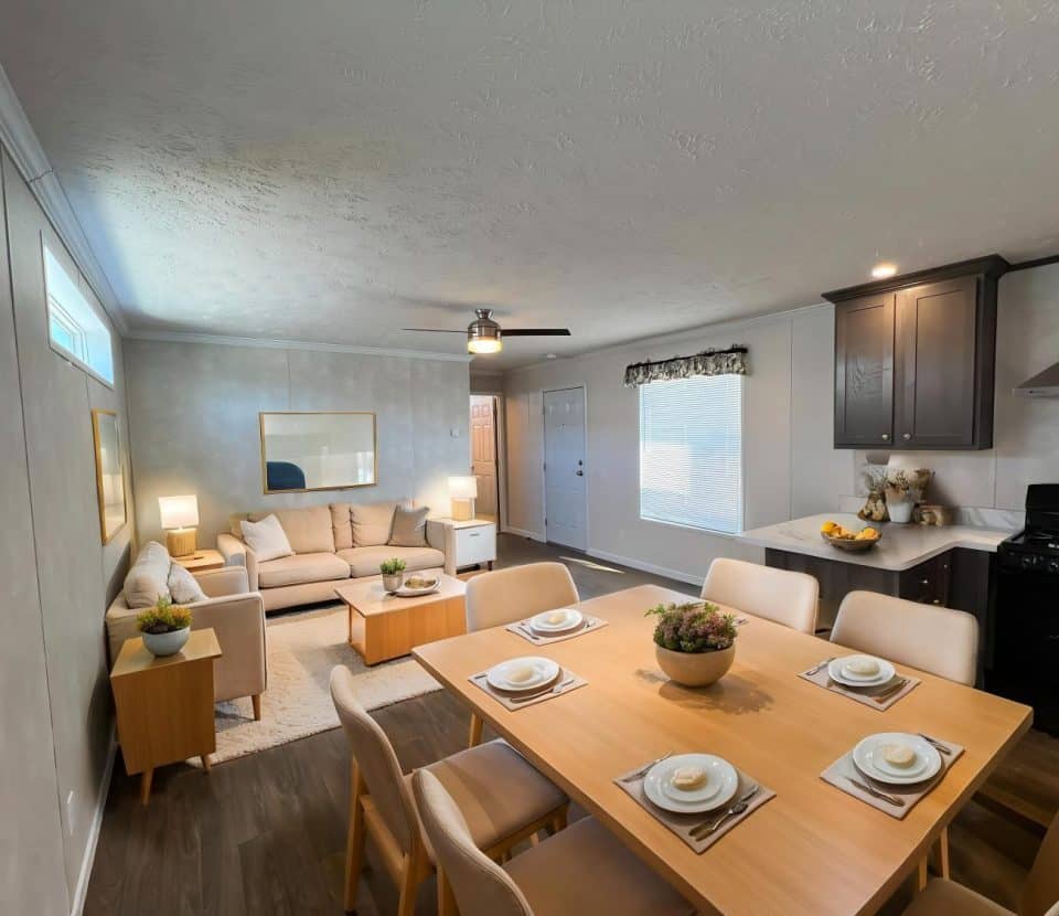 A modern dining and living area with a wooden dining table set for six, beige chairs, a cozy beige sofa, light wood furniture, and a kitchen with dark cabinets and a stove in the background.