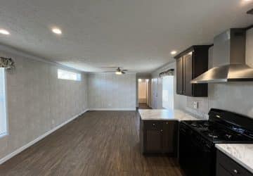 Open kitchen with dark cabinets and stainless steel appliances connects to a spacious living area with wood flooring, neutral walls, recessed lighting, and a ceiling fan. Natural light enters through windows on the left.
