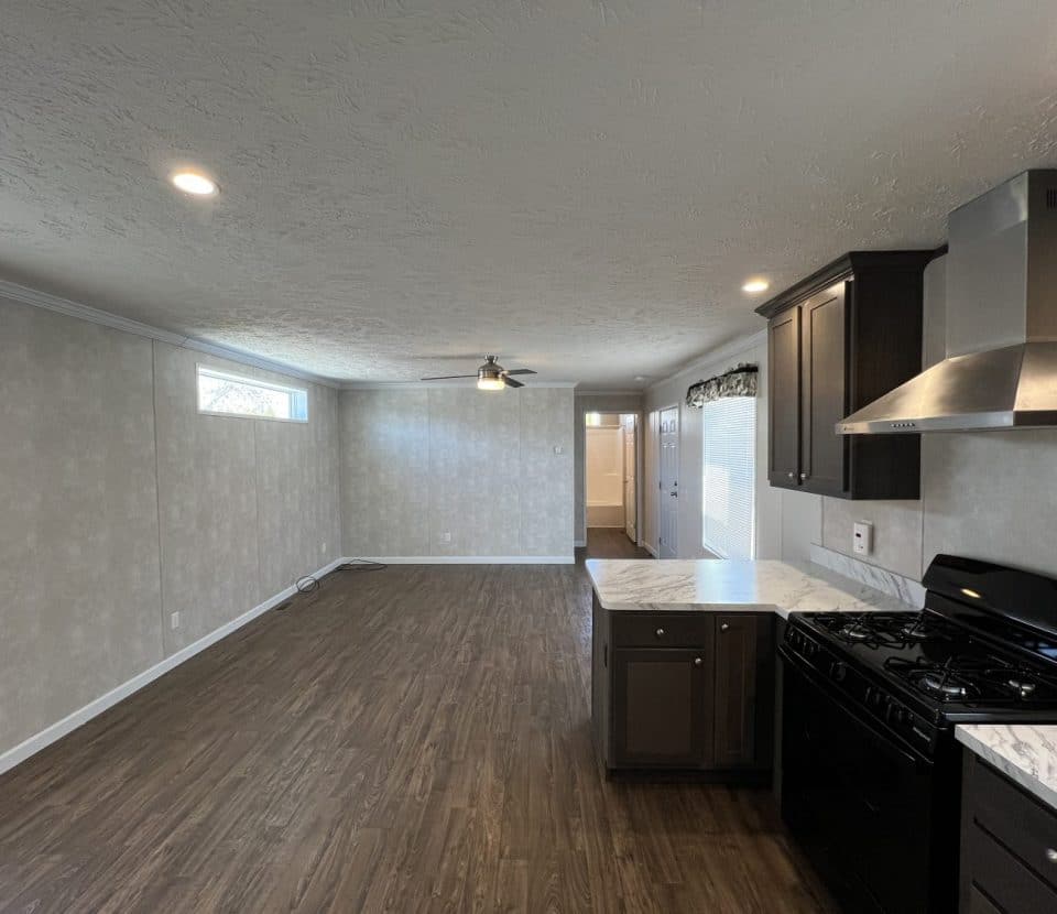 Open kitchen with dark cabinets and stainless steel appliances connects to a spacious living area with wood flooring, neutral walls, recessed lighting, and a ceiling fan. Natural light enters through windows on the left.