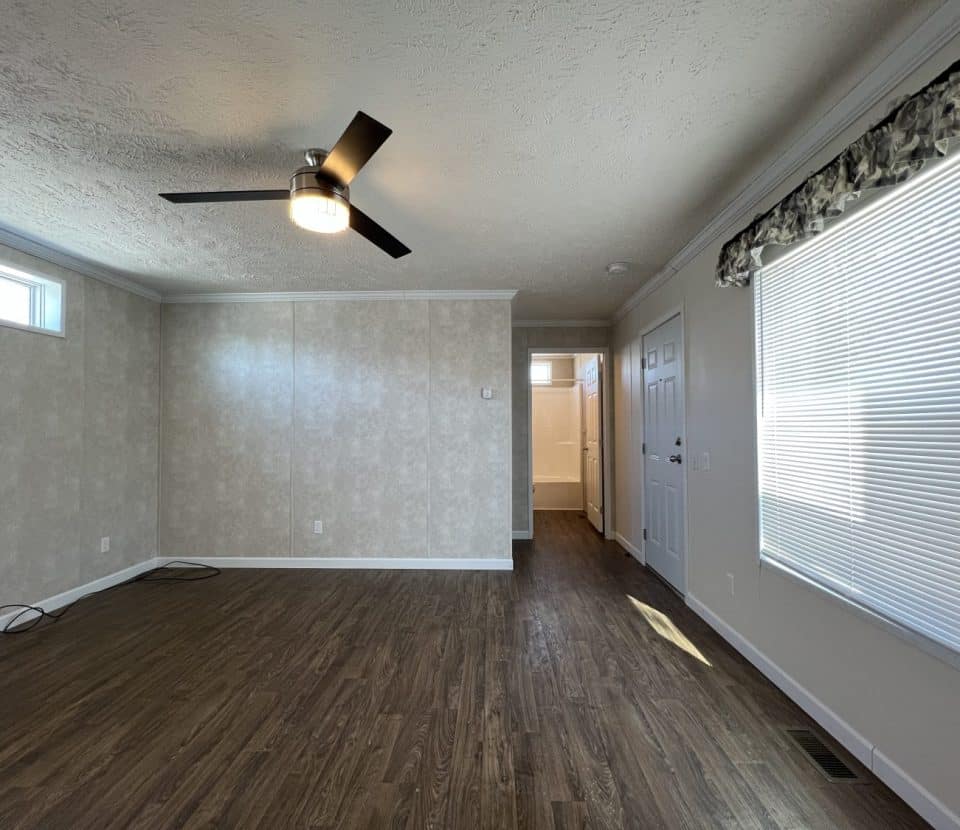 A bright, empty room with wood flooring, light-patterned walls, a ceiling fan, large window with blinds, and an open doorway leading to a hallway and another room in the background.