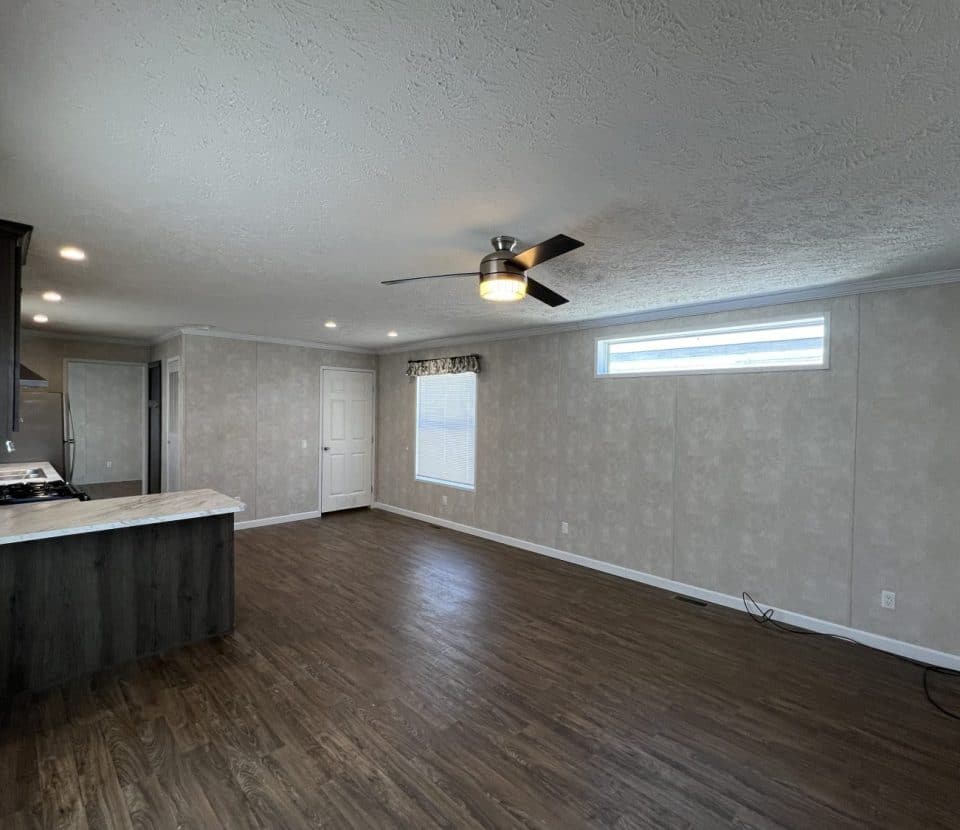 A spacious, empty living area with dark wood flooring, light textured walls, a ceiling fan with lights, a large window, and an open kitchen with dark cabinets and white countertops.