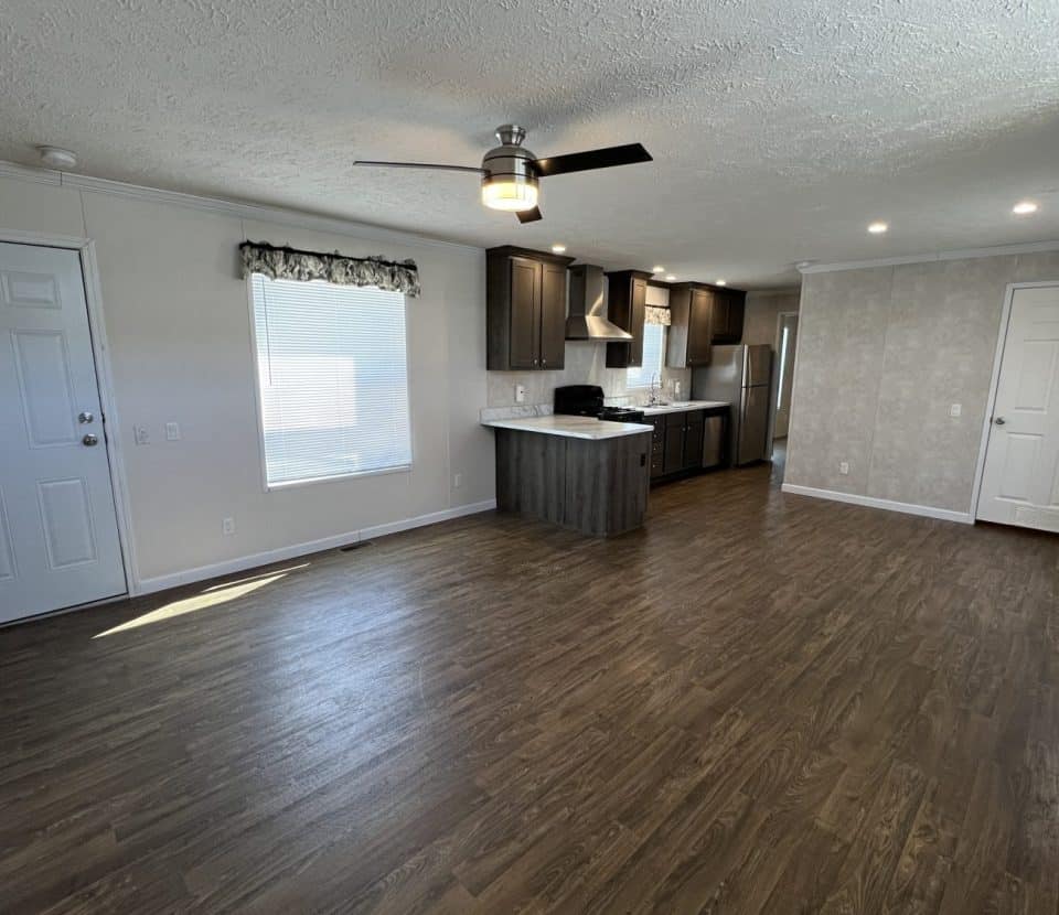 Open-concept living area with wood flooring, a ceiling fan, and recessed lighting. The modern kitchen features dark cabinets and stainless steel appliances. Light streams through a window beside the entry door.