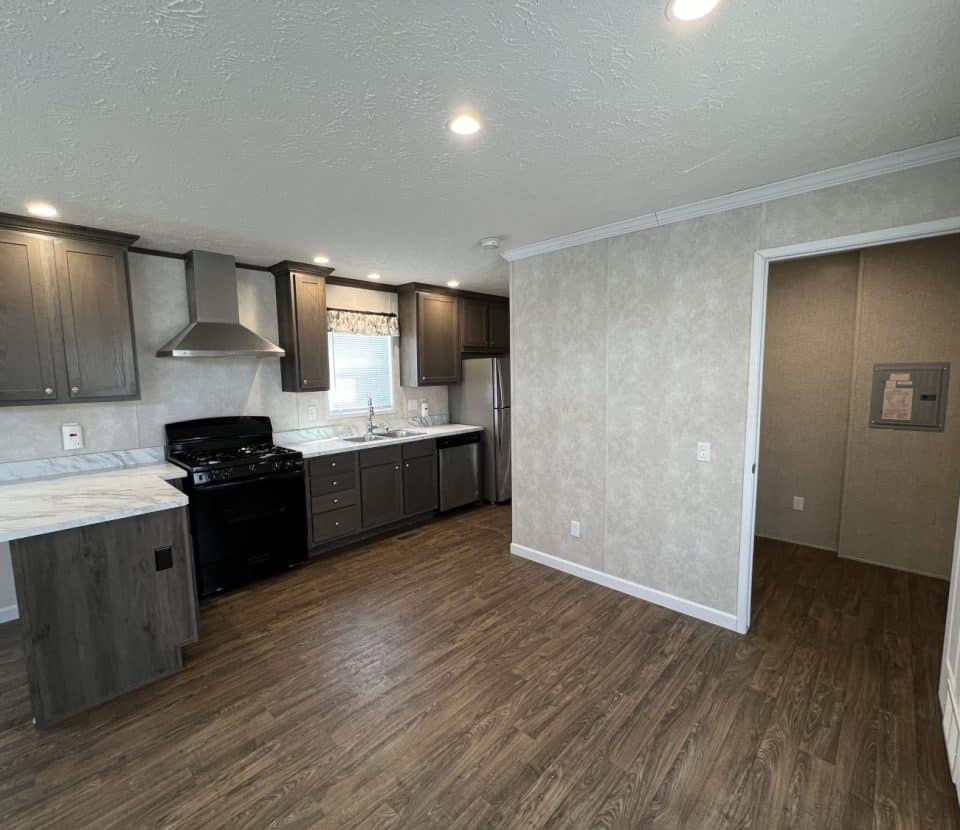 Modern kitchen with dark cabinets, stainless steel appliances, and a marble countertop. The room has wood-style flooring, recessed ceiling lights, and beige textured walls. An open doorway leads to another room.