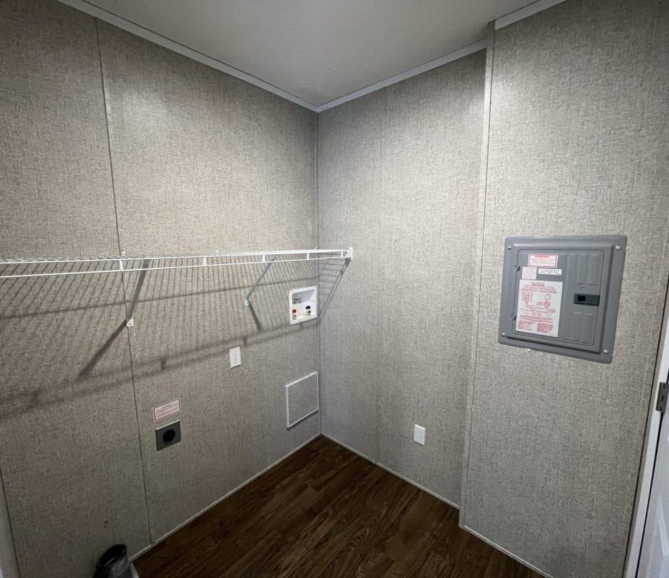 A small, empty laundry room with beige walls, a wire shelf, wall outlets, washer and dryer hookups, a fuse box, and dark wood flooring.