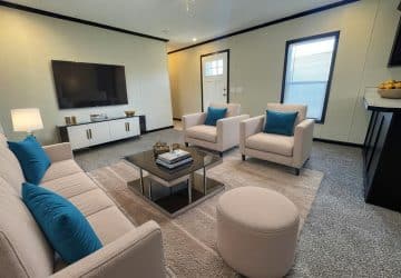 A modern living room with light-colored furniture, blue accent pillows, a coffee table with books, a flat-screen TV on a white console, a large rug, and a window and door letting in natural light.