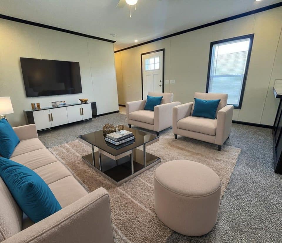 A modern living room with light-colored furniture, blue accent pillows, a coffee table with books, a flat-screen TV on a white console, a large rug, and a window and door letting in natural light.