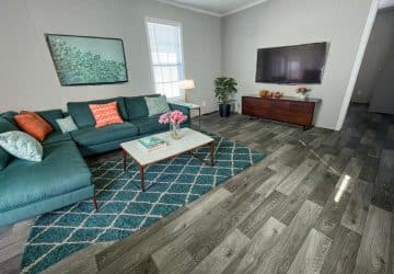 Modern living room with a teal sectional sofa, colorful pillows, a patterned rug, coffee table with flowers, TV on the wall, wooden console, potted plant, and a large window letting in natural light.