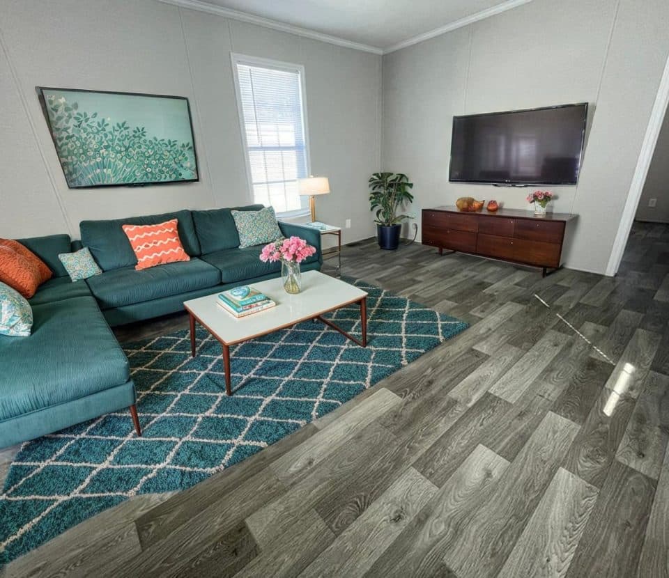Modern living room with a teal sectional sofa, colorful pillows, a patterned rug, coffee table with flowers, TV on the wall, wooden console, potted plant, and a large window letting in natural light.