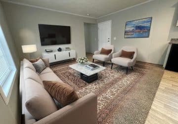 A modern living room with a beige sofa, two matching armchairs, a marble coffee table, a large rug, a TV mounted on the wall, a white media console, and a seascape painting on the wall.