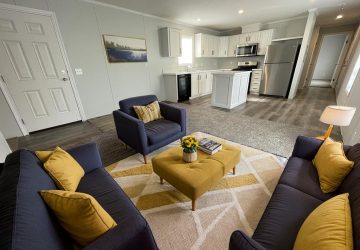 A modern open-concept living room and kitchen with navy sofas, yellow accent pillows, a matching ottoman, a patterned rug, and stainless steel appliances. A potted plant sits on the ottoman, and wall art decorates the space.
