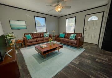 A modern living room with brown leather sofas, teal accent pillows, a wooden coffee table on a light blue rug, two large windows, a ceiling fan, and a door with a window. Decor includes flowers and framed art.