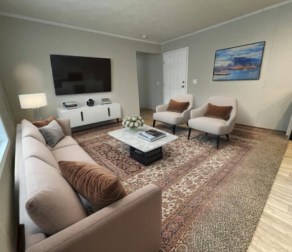 A modern living room with a beige sofa, two matching armchairs, a marble coffee table, a large rug, a TV mounted on the wall, a white media console, and a seascape painting on the wall.