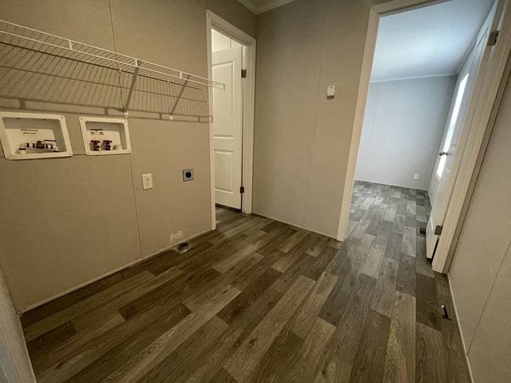 A small empty laundry room with wood-patterned flooring, white walls, a wire shelf, and hookups for a washer and dryer. Two doorways lead to adjacent rooms with similar flooring and light-colored walls.