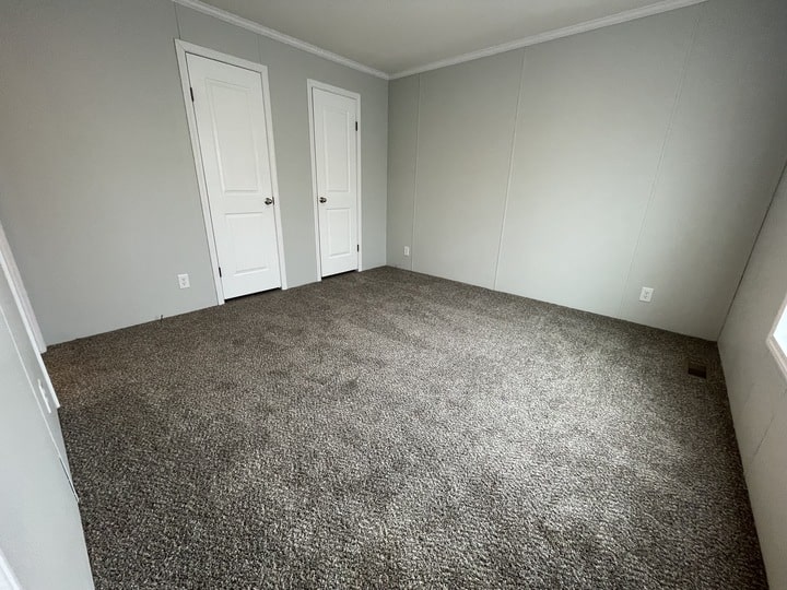Empty room with light gray walls, brown carpet, two white doors, and a window on the right side letting in natural light. The space is clean and undecorated.