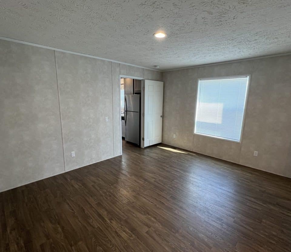 A bright, empty room with wood flooring, beige textured walls, a large window with blinds, and an open door leading to a kitchen area with a refrigerator visible.