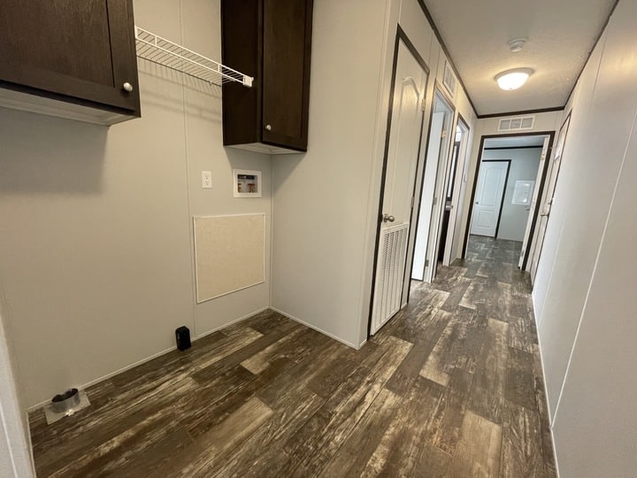 A hallway with wood-patterned flooring, white walls, and multiple doors. On the left, there is a utility area with dark cabinets, a wire shelf, and wall outlets. A ceiling light is visible at the far end of the hall.