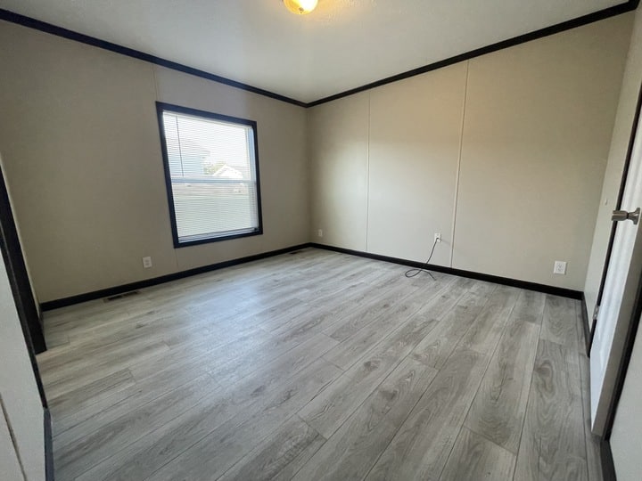 Empty room with light gray wood flooring, beige walls with black trim, a single window with blinds, a ceiling light, electrical outlets, and a visible power cord along the wall.