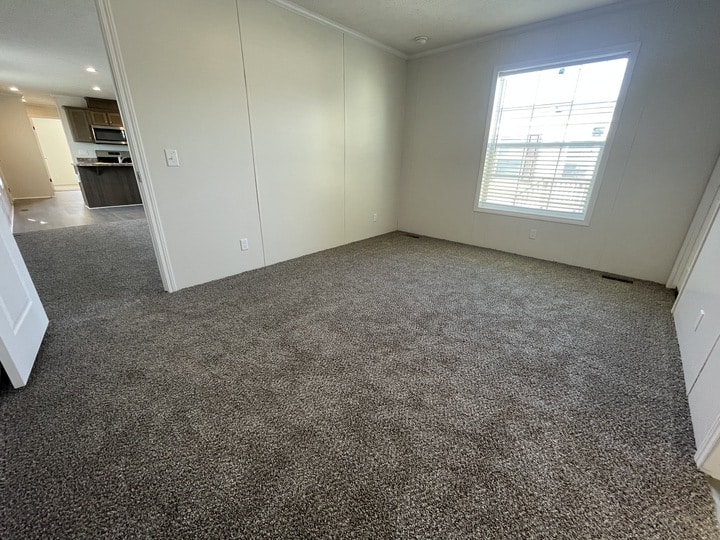 A bright, empty room with light-colored walls, gray carpet, a large window with blinds, and a view into a kitchen and dining area in the background.