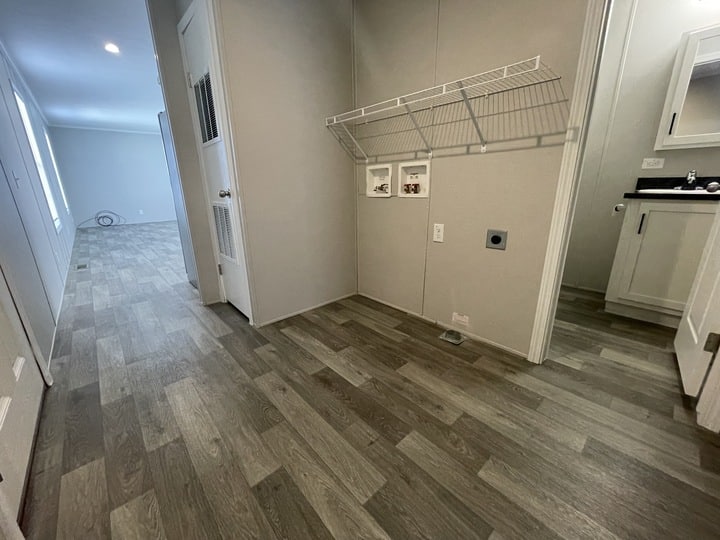 A laundry room with wood-patterned flooring, an empty white wire shelf, wall connections for a washer and dryer, and an adjacent bathroom with a white vanity and mirror. A hallway and open room are visible in the background.