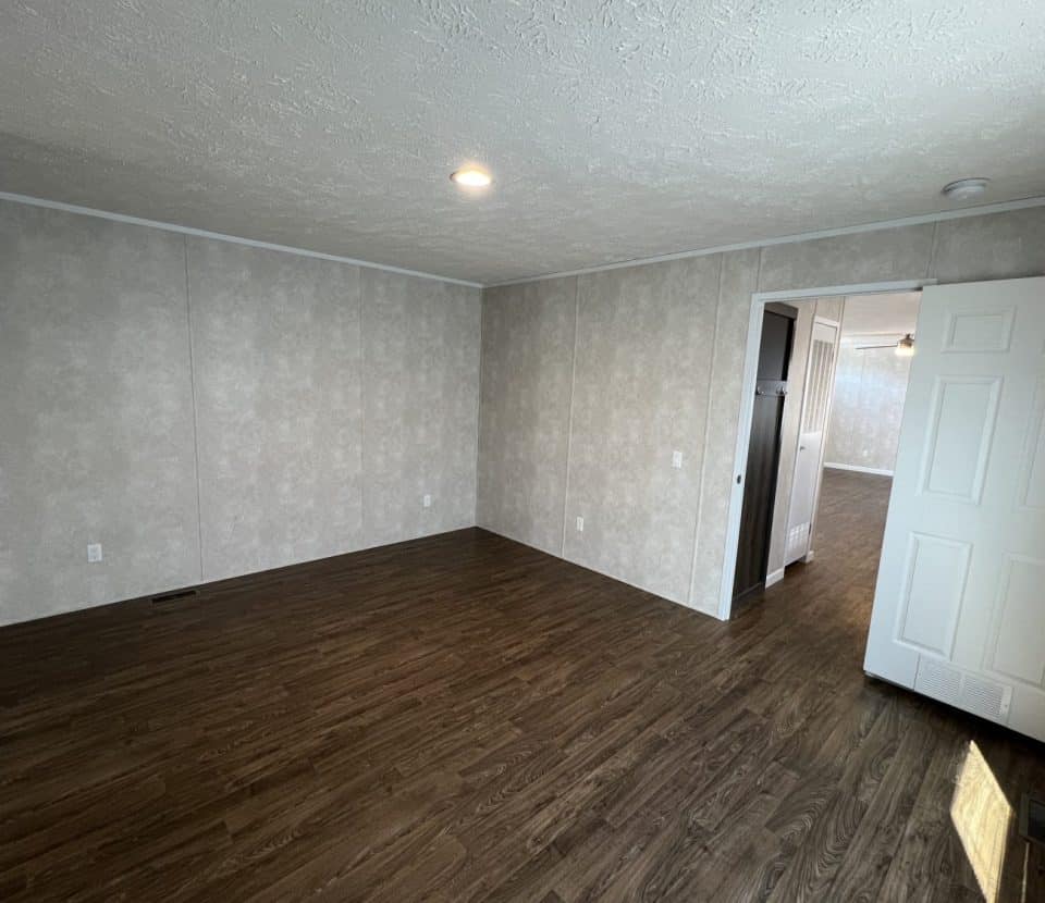Empty room with wood-style flooring, light-colored textured walls, a white ceiling with a single recessed light, and an open door leading to another bright room.