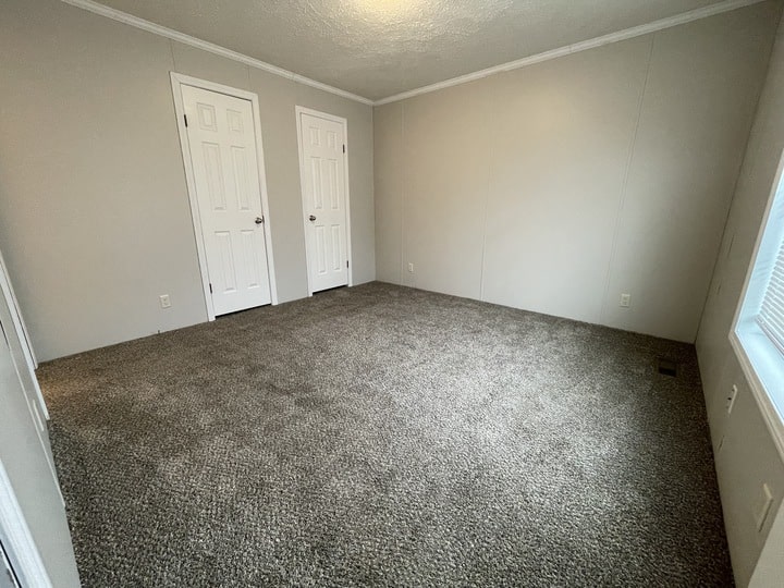 Empty room with beige walls, gray carpet, two white doors on one wall, and a window letting in natural light on the adjacent wall. The room appears clean and well-lit.