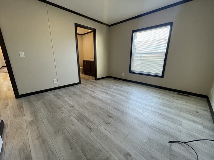 An empty room with light gray wood flooring, beige walls, dark trim, a large window with blinds, and an open doorway leading to a bathroom. A black cable is coiled on the floor near the wall.