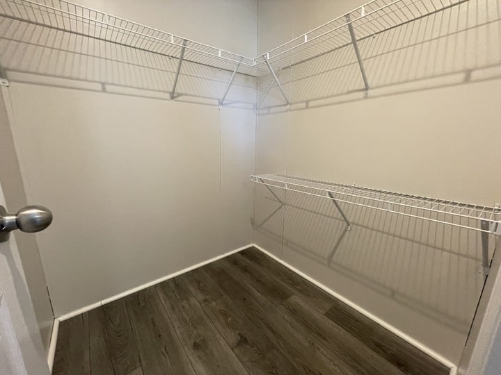 Empty walk-in closet with light-colored walls, two levels of white wire shelving, and dark wood flooring. The closet is well-lit and has a door with a silver knob partially visible on the left.