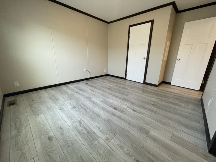 An empty room with light gray wood flooring, beige walls, white baseboards, and two white doors. There is a wall outlet and a visible loose cable near the corner.