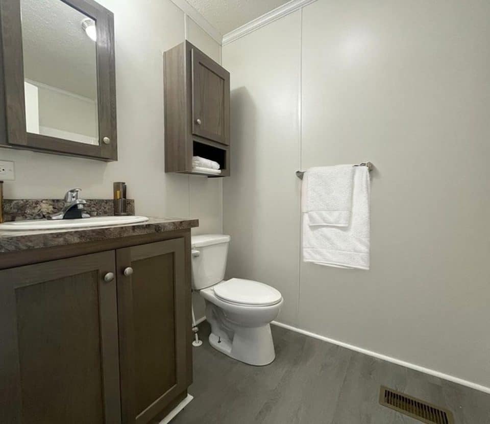 A modern bathroom with gray cabinets, a white sink, a toilet, a wall cabinet with towels, a mirror, a towel rack with a white towel, and light gray walls and flooring.