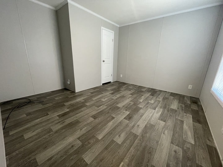 Empty room with light gray walls, white trim, a white door, and wood-look vinyl flooring in varying shades of brown. Sunlight enters from a window on the right, and electrical cords are visible in the left corner.
