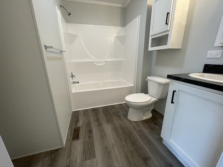Modern bathroom with wood-look flooring, a white bathtub and shower combo, a toilet, and white cabinets with black countertop, all against light gray walls.