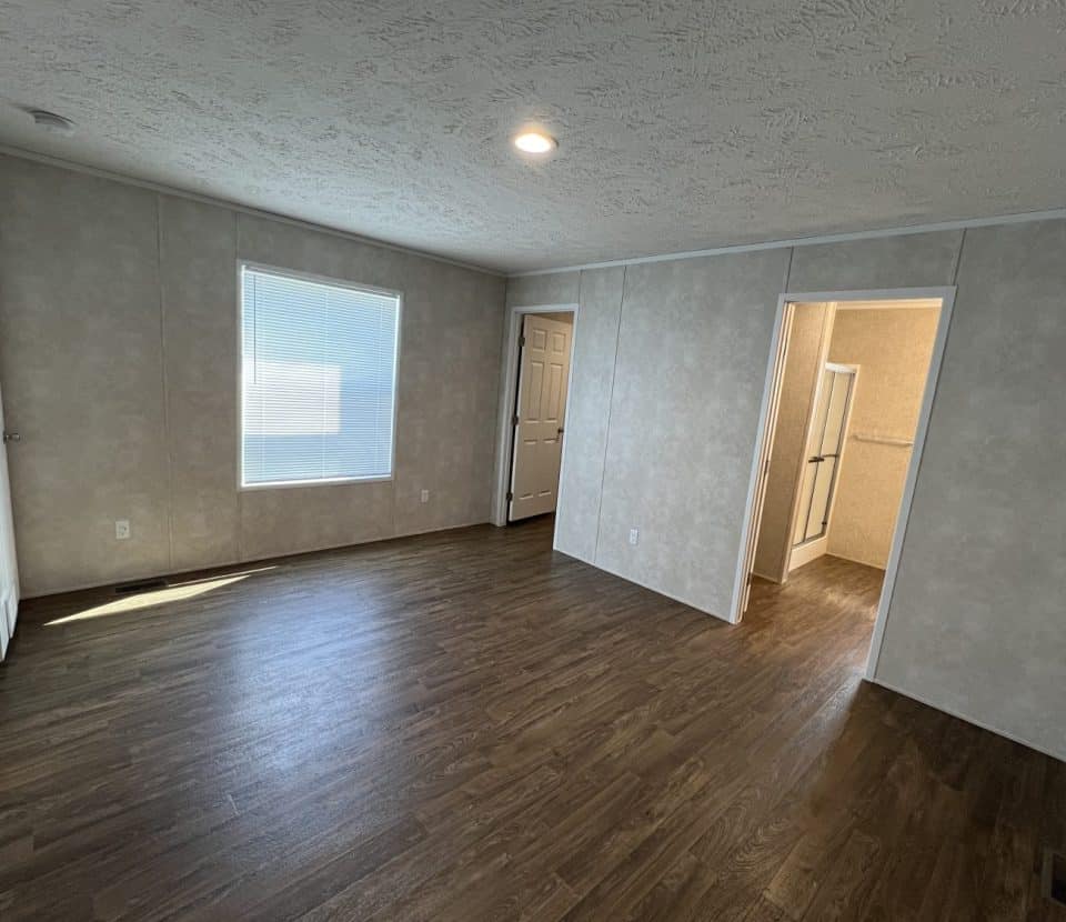 A bright, empty room with wood-look flooring, beige textured walls, a large window, and two open doorways leading to adjacent rooms. Ceiling has a recessed light and trim lines.
