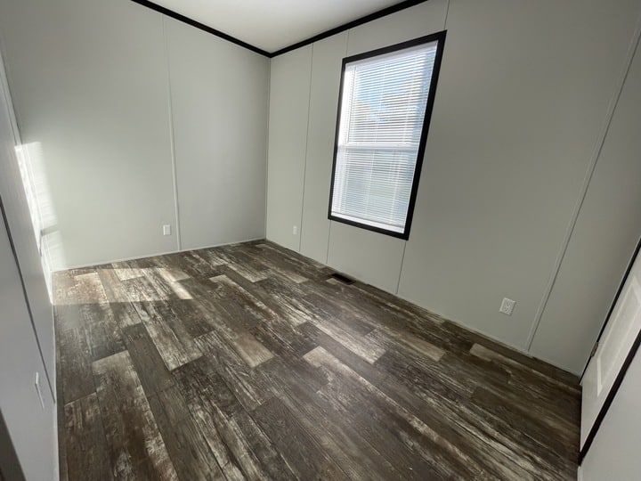 An empty room with light gray walls, a single window with blinds letting in sunlight, and dark wood-patterned flooring. The room has white trim and baseboards with visible electrical outlets.
