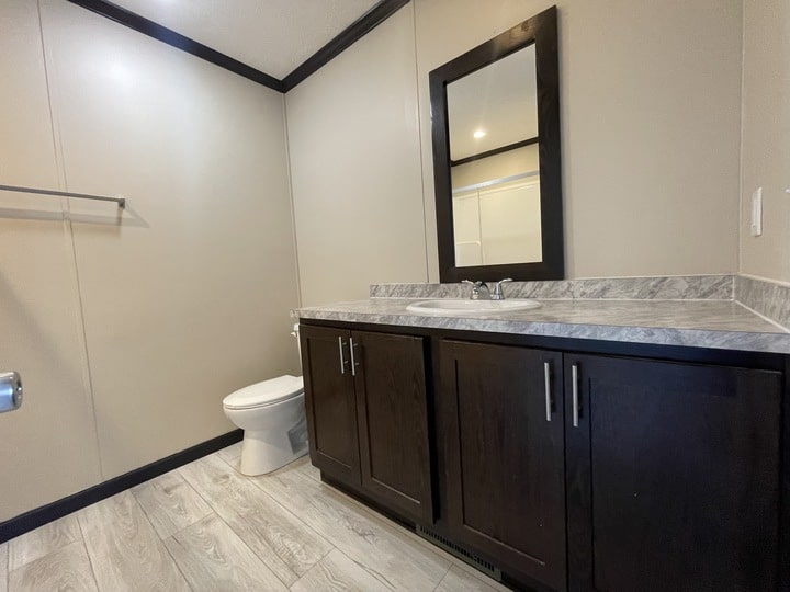 A modern bathroom with light-colored tile flooring, dark wood cabinets, a white sink with a mirror above it, and a white toilet against beige walls. A metal towel bar is mounted on the left wall.