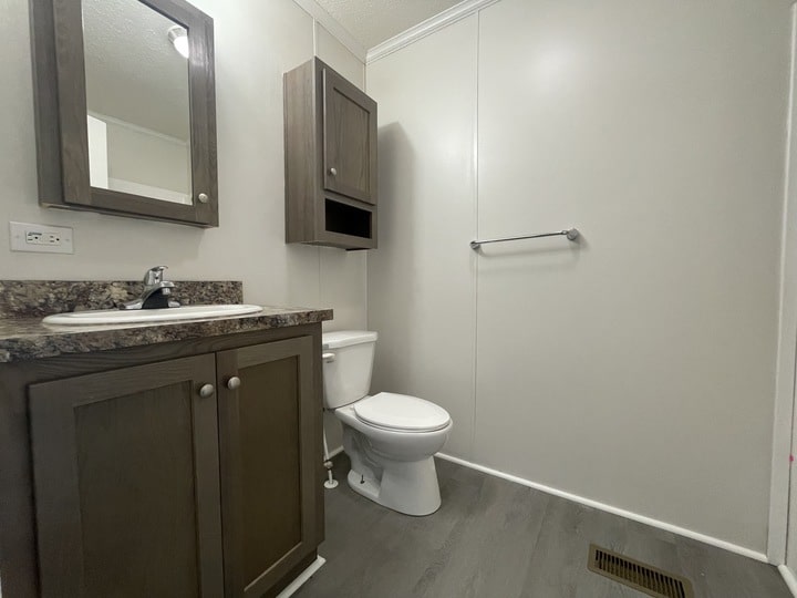A simple bathroom with a sink and cabinet on the left, a wall-mounted mirror above, a toilet in the center, a wall cabinet above the toilet, a towel bar on the right wall, and gray flooring throughout.