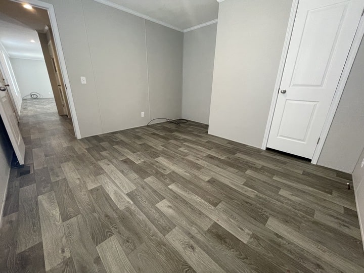 Empty room with gray wood-style vinyl flooring, light gray walls, white trim, and a closed white door. A hallway extending from the room is visible on the left side of the image.