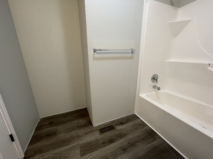 A bathroom with wood-look flooring, a white bathtub with built-in shelving, a chrome faucet, towel bar, and a wall vent on the floor. The walls are light-colored and the space appears clean and modern.