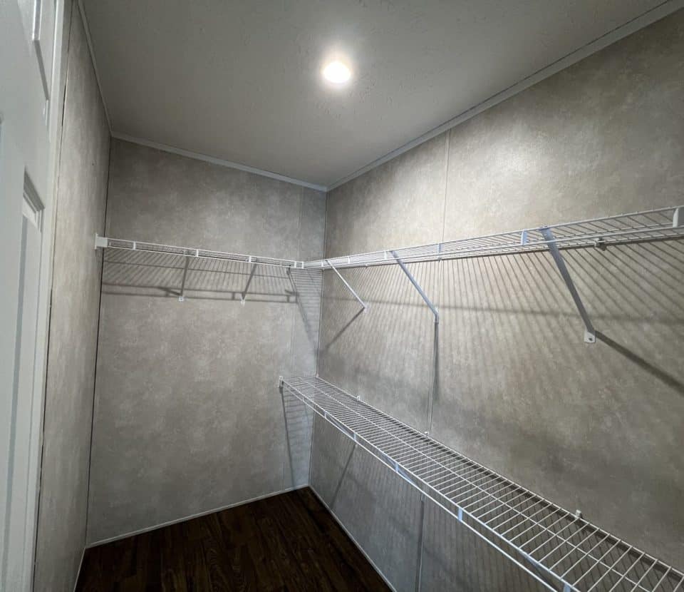 A walk-in closet with beige walls, white wire shelving on two sides, dark wood flooring, and a ceiling light centered above.