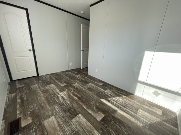 Empty room with light gray walls, two white doors, dark brown wood-patterned vinyl flooring, white trim, and sunlight streaming in through a window casting a shadow on the floor.