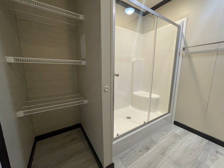 A bathroom with a glass-enclosed shower featuring a built-in seat, next to an open closet with four white wire shelves and light wood-look flooring.
