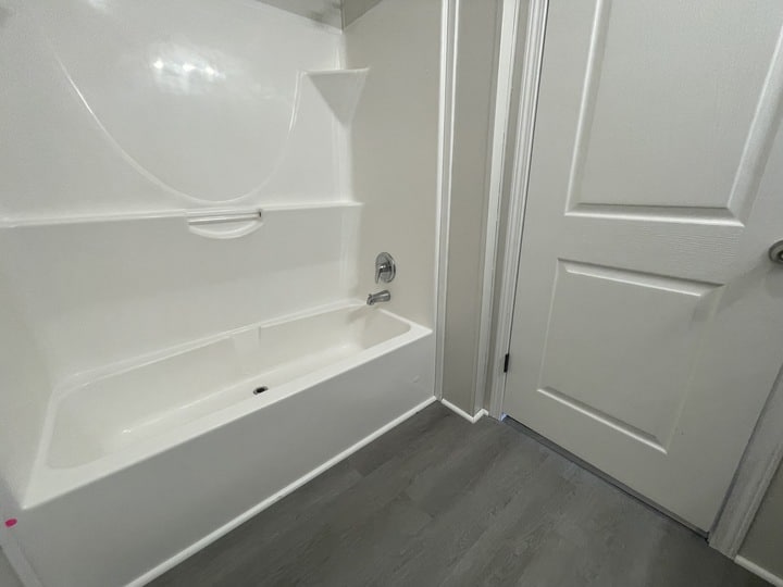 Bathroom with a white bathtub and shower combo, chrome faucet, gray paneled floor, light-colored walls, and two white paneled doors. The space is clean and minimalistic.