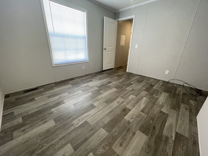 Empty room with light gray walls, a large window with closed blinds, wood-patterned vinyl flooring, an open white door, and a few visible electrical outlets.