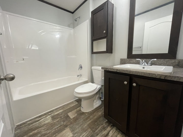 A modern bathroom with a white bathtub-shower combo, toilet, and a dark wood vanity with a sink and mirror. The floor has a wood-look pattern, and there are matching dark wood cabinets above the toilet and under the sink.