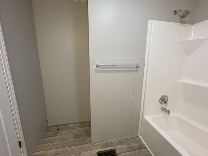 A bathroom with light wood flooring, a bathtub and shower on the right, a silver towel rack on a plain wall, and a floor vent in front of the tub. The walls are white and the space looks clean and minimal.