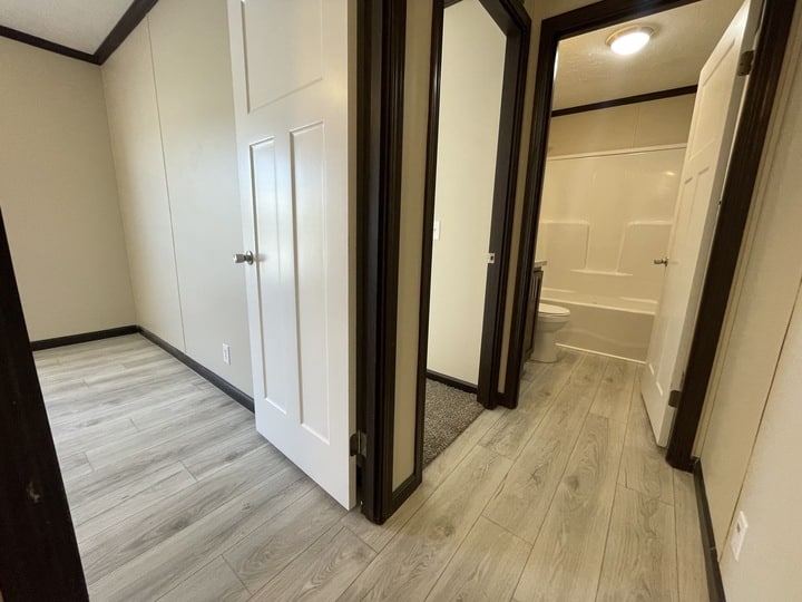A hallway with light wood flooring leads to a bathroom with a white bathtub and toilet on the right, and an empty room with the same flooring on the left. The walls are beige with dark trim and white doors.