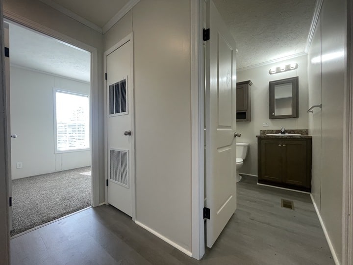 A hallway with gray flooring leads to a bathroom with a vanity, mirror, and toilet on the right. An open door on the left shows a carpeted room with a large window letting in natural light.
