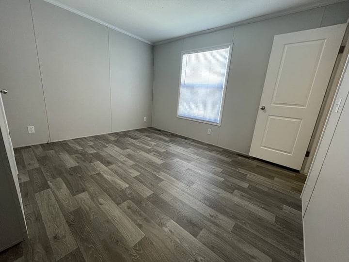 Empty room with light gray walls, a window with blinds, wood-patterned vinyl flooring, and a white door slightly open. The room is bright and unfurnished.