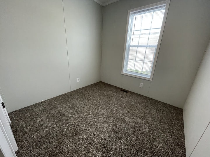 A small empty room with light-colored walls, a large window with blinds, and brown carpet flooring. Sunlight is coming through the window, creating a bright and clean appearance.