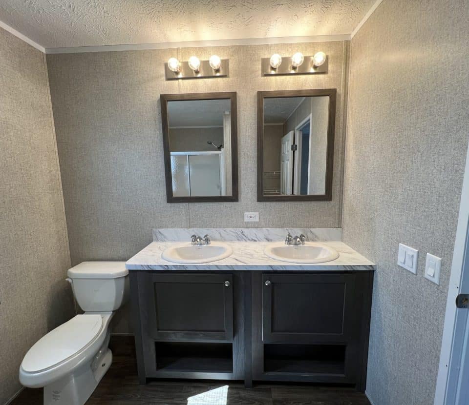 Modern bathroom with two sinks in a marble countertop, dark cabinets, two mirrors above, two light fixtures with three bulbs each, a toilet to the left, and a partial view of a shower and doorway to the right.